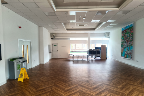 The main hall is a large rectangular space with wooden flooring and windows at one end. There is a piece of art hanging on the wall on the left wall and double doors on the right wall.
